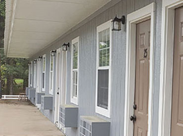Outside of motel rooms with light blue walls, white trim around window and door frames, and brown doors next to a wooded area with a white, picket fence